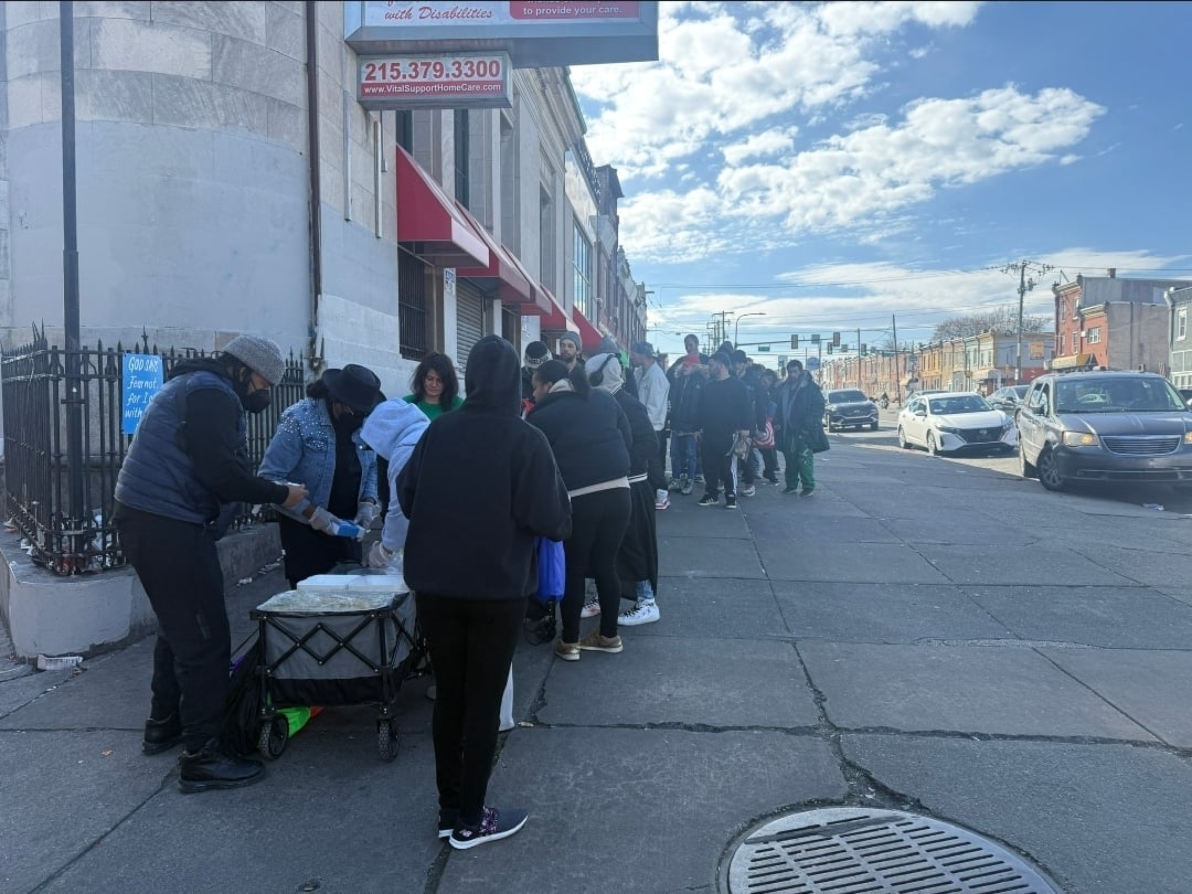 People standing in line on a city sidewalk outside a storefront on a sunny day