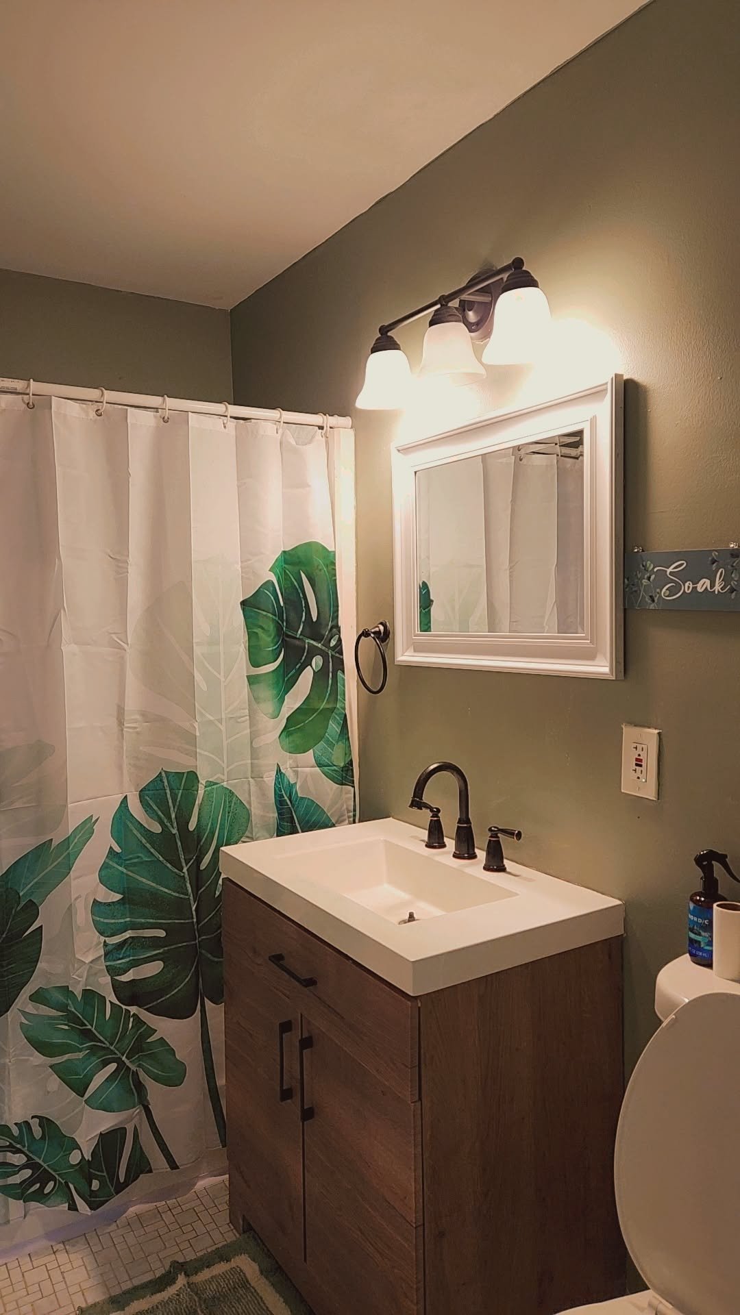 Modern bathroom with monstera leaf shower curtain, wooden vanity, white sink, framed mirror, and bronze fixtures